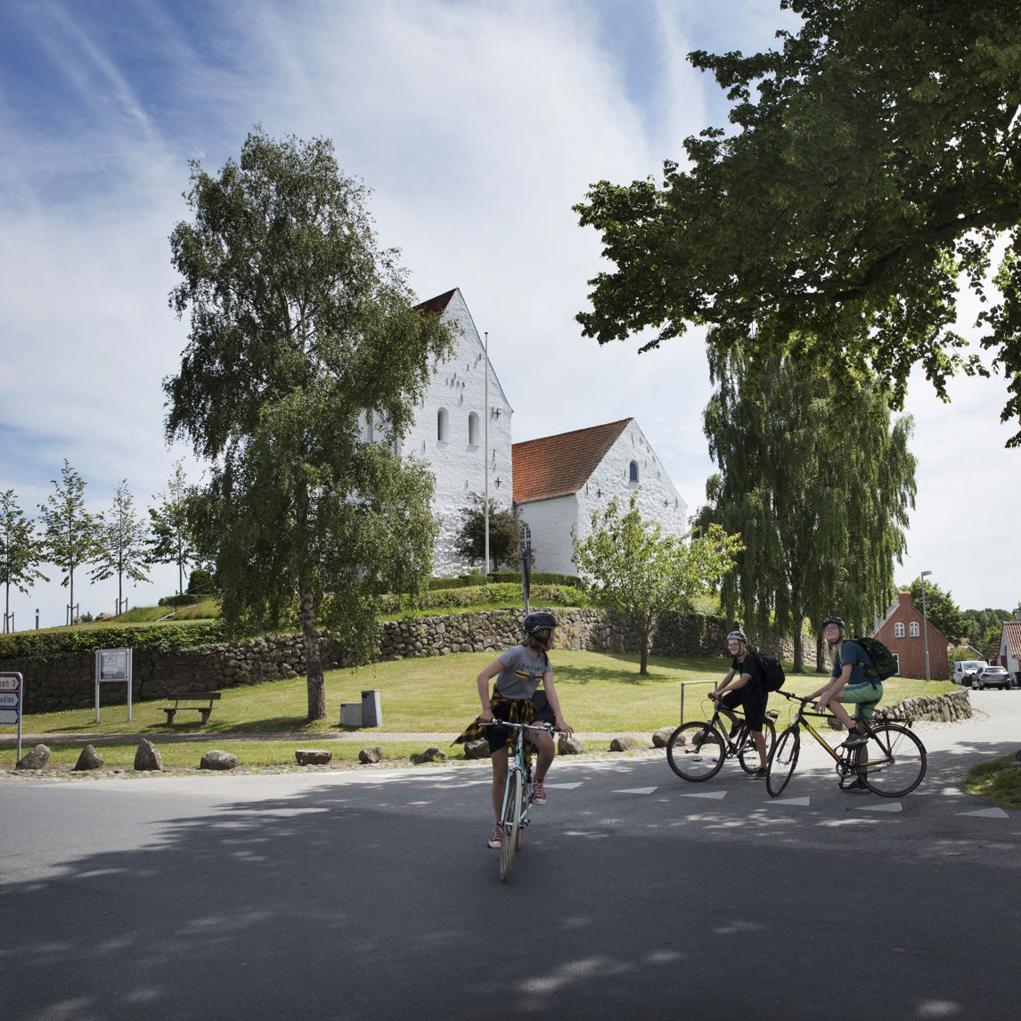 tre cykelister foran hvid kirke i grønne omgivelser